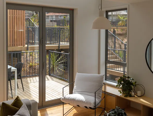 A bright, compact living room with a pale upholstered sofa, patterned and olive cushions, a black rectangular coffee table, a white lounge chair, and a low timber console. Full-height glazed doors and windows open onto a timber deck and metal-railed exterior walkway, bringing in strong natural light. The room has light timber flooring, white walls, a neutral rug, a simple pendant light, and indoor plants.