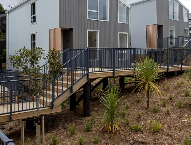 Exterior view of a contemporary multi-unit residential development on a sloped, landscaped site. Two compact gabled buildings feature white and dark grey vertical cladding, large rectangular windows, timber entry accents, and elevated timber decks connected by dark metal balustrades and stairs.