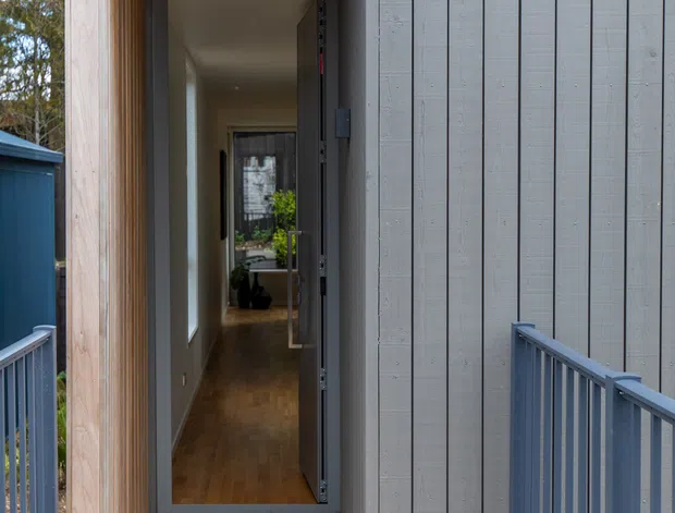 A narrow exterior walkway and timber deck lead to an open front door set within a deep recessed entry. The building is clad in vertical grey boards, with natural timber lining the entry reveal and decking, and blue-grey metal railings along the elevated path. Through the doorway, a long interior corridor with timber flooring and a glimpse of plants and furnishings is visible. The image has a clean, contemporary architectural character.