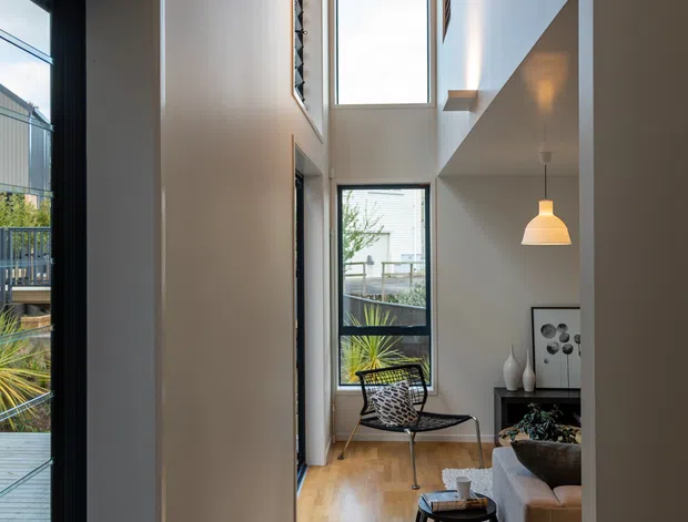 A narrow, double-height residential living area viewed from an adjoining hallway or entry. The space features warm timber flooring and a timber-lined ceiling, white walls, tall black-framed windows, a small seating area with a sculptural lounge chair and sofa, a round side table, pendant lighting, and built-in shelving. An outdoor deck and planted garden are visible through the glazing.