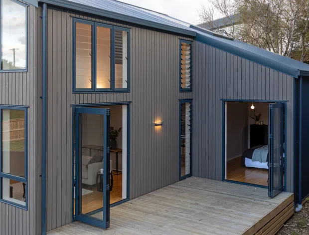 Exterior view of a compact modern two-storey dwelling clad in dark vertical board-and-batten-style siding, with dark-framed windows and glazed doors opening onto a timber deck. The recessed courtyard-like deck connects indoor living and bedroom areas, and the sloping roof and clean linear form create a contemporary architectural character.