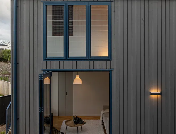 Exterior view of a compact contemporary two-storey dwelling with dark vertical cladding, a corrugated metal roof, dark-framed windows, and a timber deck. A large open glazed door reveals a softly lit living room with timber flooring, a pale rug, pendant lighting, and seating. The facade also features discreet wall lighting, external downpipes, a satellite dish, and rooftop antenna.