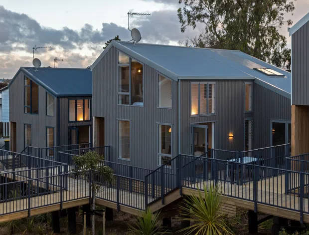 Exterior view of a contemporary multi-unit residential development with dark grey vertical metal cladding, pale grey pitched metal roofs, extensive glazing, and elevated timber walkways with dark metal balustrades. The buildings sit above a landscaped, lightly planted site with native-looking vegetation, viewed at dusk beneath a cloudy sky.
