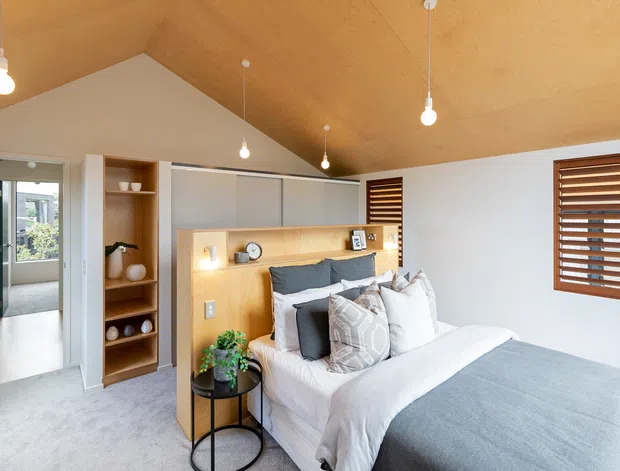Bright, compact bedroom with a pitched plywood-lined ceiling, white walls, soft grey carpet and a neatly made bed. Built-in timber shelving and a timber headboard feature integrated bedside lighting and display niches. Warm pendant bulbs, timber venetian shutters and neutral grey-and-white bedding create a calm contemporary interior.