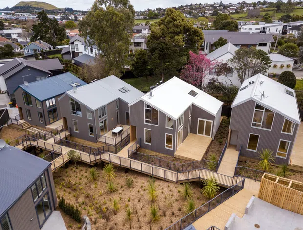 Aerial view of a contemporary multi-home residential development on a sloping suburban site. Several compact detached homes feature dark vertical cladding, light metal gable roofs, large windows, and interconnected timber decks and walkways. Native-style landscaping surrounds the homes, with established trees and a dense suburban neighbourhood visible beyond.