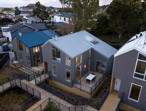 Elevated exterior view of a contemporary multi-building residential development on a sloped suburban site. Several compact homes feature dark grey vertical cladding, pitched metal roofs, timber decks, extensive connecting timber walkways and dark metal balustrades. The homes overlook a leafy neighbourhood with a low hill in the distance.