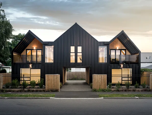 Front exterior view of a contemporary multi-unit residential building with three steeply gabled volumes clad in dark vertical metal, glazed upper-level balconies, warm interior lighting, timber fencing, and a central covered pedestrian passage. The building sits directly along a suburban street beneath an overcast sky.