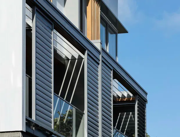 A close-up exterior view of a contemporary multi-storey residential building with crisp geometric forms, dark horizontal cladding, pale vertically ribbed panels, timber-look vertical screening, projecting eaves, and large glazed openings. A dark timber fence occupies the foreground, with blue sky and limited planting in the background.