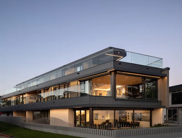 Exterior view of a contemporary multi-level residential building at dusk, featuring dark horizontal cladding, extensive floor-to-ceiling glazing, glass balustrades, projecting balconies, recessed warm lighting, and a rooftop terrace. Interior living and dining areas are visible through the windows, with a neighbouring dwelling partially visible at right.
