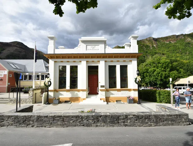 Street-level view of a small historic civic building with a symmetrical white-painted facade, classical pilasters, decorative cornice, tall windows, a central dark timber door, and a raised stone terrace. A nearby museum building, landscaped hedges, dramatic hills, and cloudy sky are visible in the surroundings.
