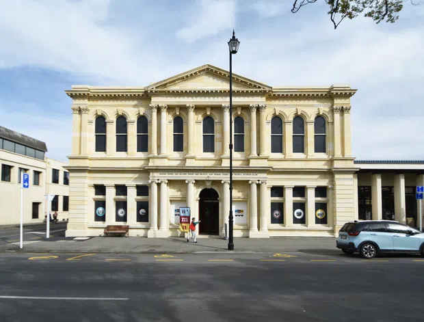 Street-facing view of a grand historic neoclassical civic or commercial building with a symmetrical cream stone or rendered facade, classical columns and pilasters, an ornate entablature, triangular pediment, and tall arched windows. The building fronts an urban street with adjacent modern structures and street parking.