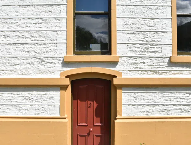 Close-up exterior view of a traditional masonry facade with heavily textured white-painted rendered walls, warm ochre trim and lower wall panels, timber-framed windows, and a dark red paneled entrance door beneath a shallow arched hood. The image focuses on the building frontage and architectural detailing.