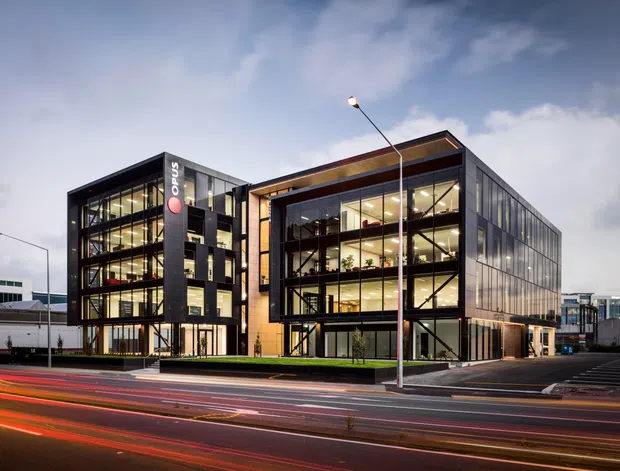 Exterior architectural photograph of a contemporary multi-storey commercial office building at dusk. The building has extensive floor-to-ceiling glazing, dark structural framing, a prominent recessed entrance volume with warm timber-toned cladding, and an adjacent darker office wing with visible interior workspaces. It fronts a busy urban road with light trails, landscaped strips, and surrounding commercial buildings.