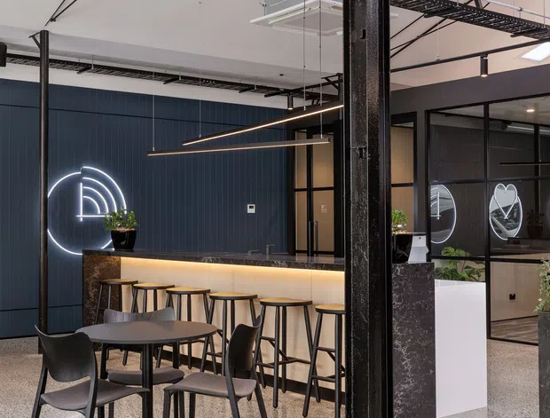 A contemporary commercial café or breakout area within an industrial-style office interior. The space features a long illuminated bar with stools, small dining tables and chairs, dark vertical wall panelling, black steel structural columns, exposed cable trays and services, suspended linear lighting, internal glazed meeting-room partitions, and a polished speckled concrete or terrazzo floor. A neon-style circular sign and indoor plants add visual accents.