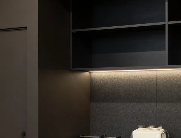 A dark, refined built-in workspace or office nook with black open shelving above a low black cabinet, integrated warm under-shelf lighting, and a textured grey wall panel behind the work surface. Office equipment sits on the cabinet, with a simple flush door at left.