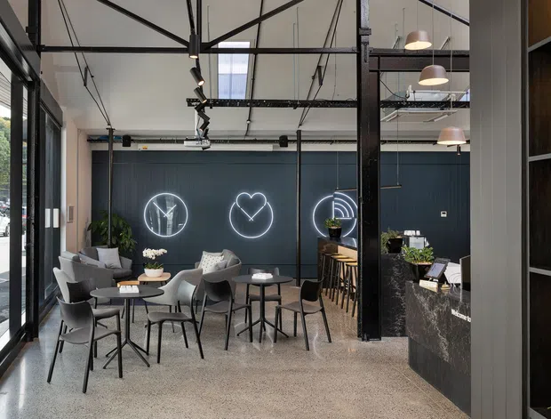 A contemporary commercial café or hospitality interior with an open-plan seating area, polished concrete floor, dark charcoal feature wall with illuminated graphic signage, exposed black structural framing and services, pendant lighting, indoor plants, and a stone-clad service counter with timber shelving. Full-height glazing opens the space visually to the street.