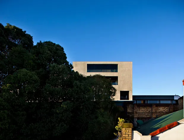 Exterior architectural view of a contemporary multi-level building, partially screened by mature trees. The main structure has a simple rectangular form with pale rendered or masonry walls, broad dark-framed horizontal windows, and a flat roof. Lower adjoining volumes feature dark glazing, exposed brickwork, and a sloping green metal roof with red trim. Clear blue sky forms the background.