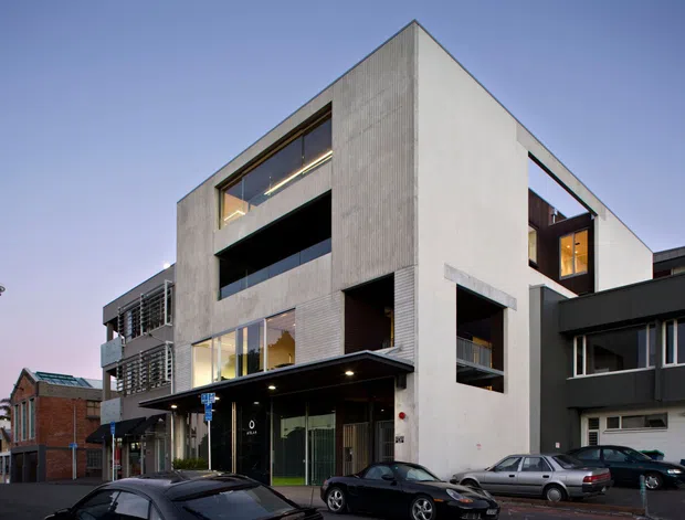 Street-level exterior view of a contemporary multi-storey urban building with an angular flat-roofed form, pale concrete or rendered walls, dark recessed openings, extensive glazing, balconies, and ground-floor commercial or entry frontage. Adjacent older brick buildings and a prominent city tower are visible in the background.