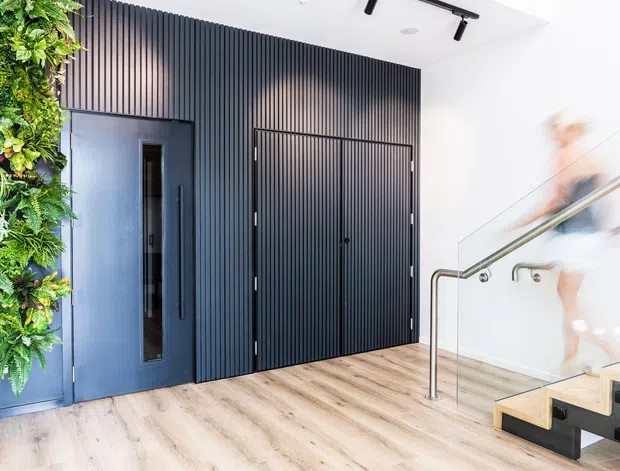 Bright contemporary entryway or landing with pale timber-look flooring, a dramatic black vertically ribbed feature wall and concealed double doors, a dark blue-grey door, a lush vertical garden wall, and a modern stair with glass balustrade and brushed metal handrail. A blurred person is moving on the stairs.
