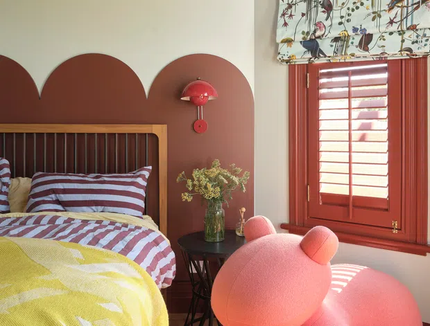 A colourful, playful bedroom featuring a timber bed with striped bedding, a terracotta scalloped painted feature wall, matching red-toned timber window and shutters, and a patterned botanical-and-bird roller blind. A large pink sculptural toy-like chair or seating piece dominates the foreground, with a small black side table and vase beside the bed. Timber flooring and strong sunlight create a warm, cheerful interior.