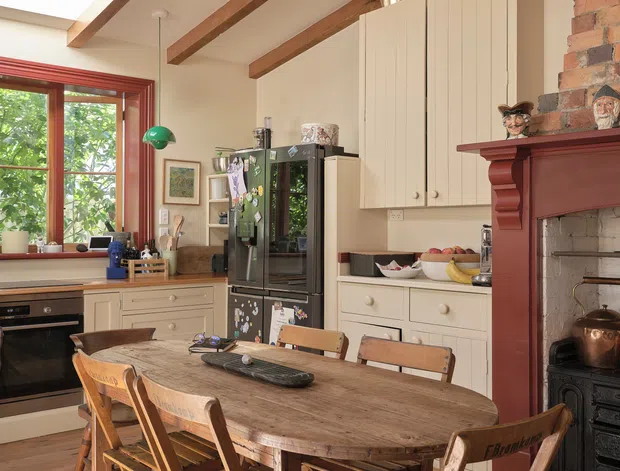 A warm, traditional kitchen-dining space with cream cabinetry, a large rustic timber dining table, folding timber chairs, exposed timber ceiling beams, and a red-painted brick fireplace with a dark cast-iron stove. The room includes a black refrigerator, timber flooring, a red-trimmed window, and a brass pendant light. The overall character is farmhouse-inspired and lived-in.