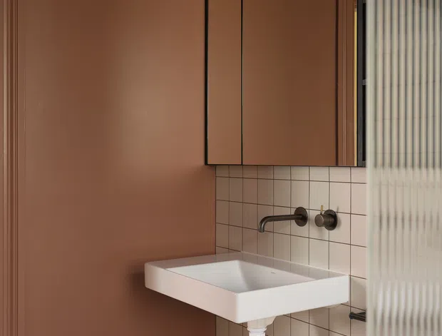 Compact contemporary bathroom vanity with a wall-mounted white rectangular basin, exposed dark metal plumbing, wall-mounted faucet, dusty terracotta painted walls, pale square tiled splashback, and terrazzo-style lower wall tiles. A mirrored cabinet with globe lighting is mounted above the basin, while ribbed glass partially screens the right side.