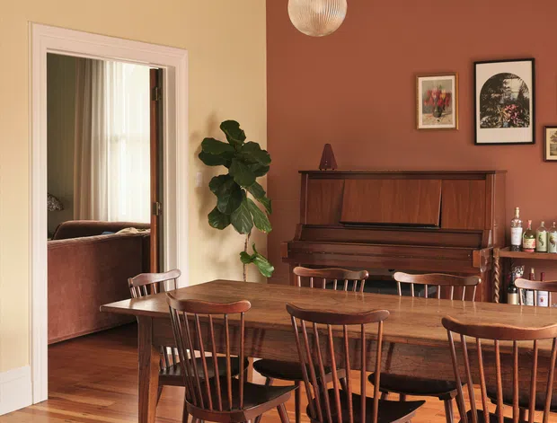 Warmly styled dining room with a long solid-wood dining table and matching spindle-back wooden chairs, an upright piano against a terracotta feature wall, framed artwork, a bar cabinet, a pendant light, and decorative painted ceiling mouldings. Timber flooring extends toward a connected living room visible through the doorway.