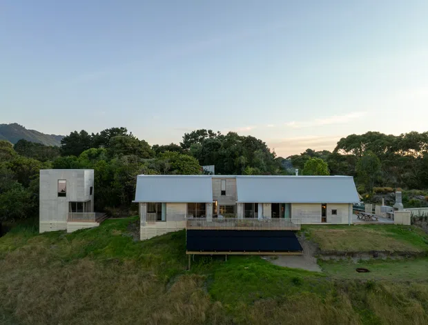 A wide exterior view of a contemporary single-storey residential home set within a leafy, semi-rural landscape. The house has a long low-profile form with a pale corrugated metal pitched roof, light timber or pale cladding, extensive glazing, and a separate cubic two-storey wing on the left. Decking and outdoor paving sit in front of the dwelling, with a dark rectangular structure in the foreground.