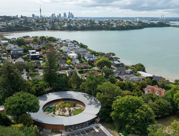 Aerial view of a leafy suburban waterfront setting with a distinctive circular, low-rise building in the foreground, surrounded by residential neighbourhoods, dense greenery, and a broad harbour or inlet. A high-rise city skyline and bridge are visible in the distance.