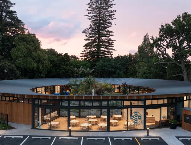 Exterior view of a low, circular or oval-shaped contemporary building set within mature landscaping at dusk. The building has a broad flat roof with a large central oval opening, extensive floor-to-ceiling glazing, dark metal window frames, and warm timber-clad wall sections. A brightly lit communal interior with tables and seating is visible through the glazing, while marked parking spaces occupy the foreground.