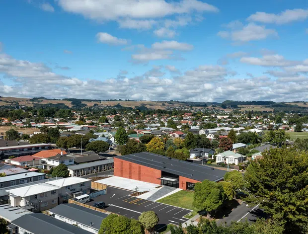 Aerial view of a suburban townscape with a modern low-rise brick building and dark pitched roof in the foreground, surrounded by residential and institutional buildings, paved parking areas, mature trees, and rolling hills under a partly cloudy blue sky.