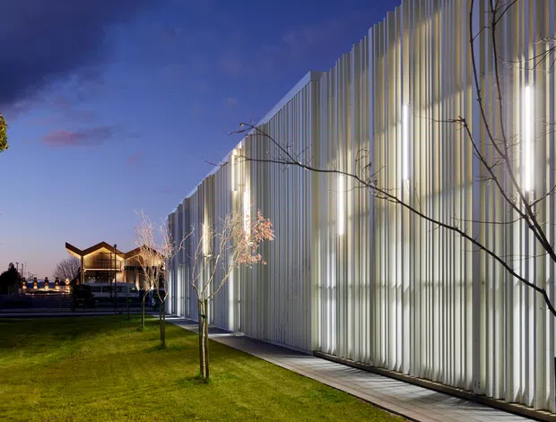 Twilight exterior view of a long contemporary building wrapped in vertically ribbed, translucent light-coloured cladding. The illuminated facade creates a strong linear rhythm, with a narrow paved path and manicured lawn in the foreground, young trees, and a dark blue evening sky. A small lit pavilion-like structure is visible in the distance.