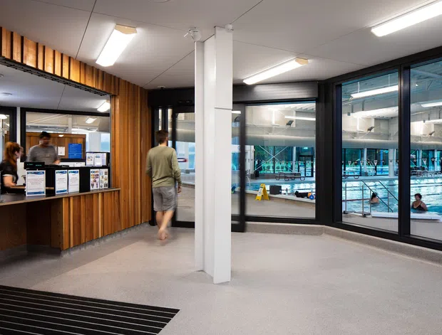 Interior view of a modern aquatic centre reception and circulation area. A timber-lined reception counter sits to the left, while large black-framed glazed partitions provide views into an indoor swimming pool. The space has a light grey carpeted floor, white ceiling panels, exposed white structural columns, and linear fluorescent lighting. People and pool activity are visible beyond the glazing.