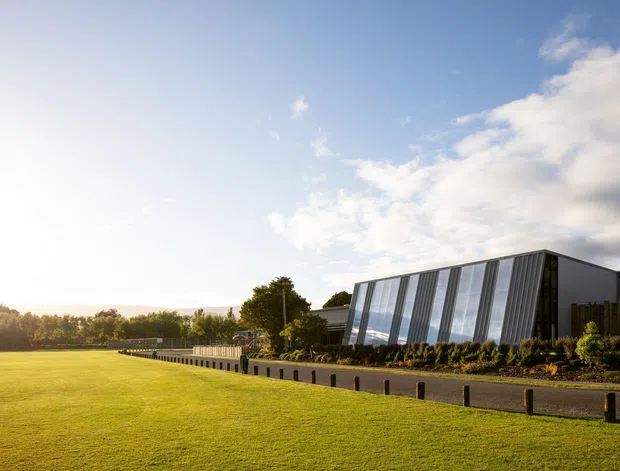 Exterior architectural photograph of a contemporary, single-storey building set beside a broad manicured lawn. The structure has a strongly angled facade with repeated tall glazed panels, dark metal framing and cladding, and landscaped planting along a paved approach. The image is captured in warm low-angle sunlight with a blue, partly cloudy sky.