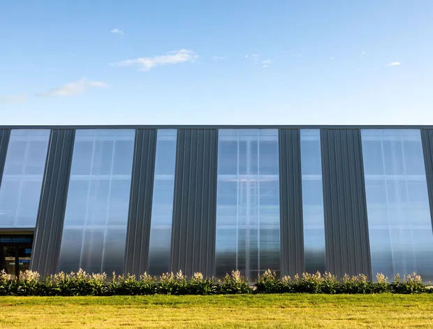 A low, modern commercial-looking building facade viewed from the lawn. The elevation is composed of dark vertically ribbed cladding with alternating full-height glazed panels and narrow solid fins. Reflections of the sky and surrounding landscape are visible in the glazing, with a planted border along the base and a clear blue sky above.