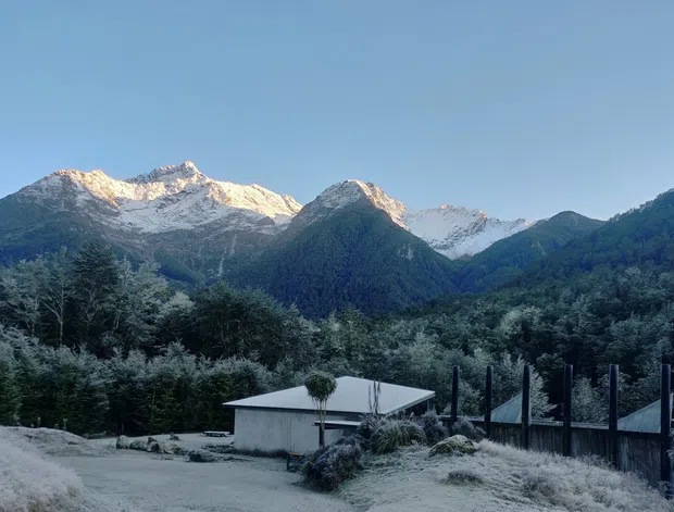 Exterior view of a simple low-profile building set within a forested mountain landscape. Snow-dusted alpine peaks dominate the background, while a gravel approach, corrugated metal-roofed structure, timber fencing, and landscaped planting occupy the foreground. The scene has a cool, tranquil, remote character.