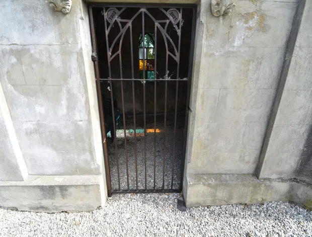 Close-up exterior view of a narrow decorative wrought-iron gate set within a weathered pale stone or plaster facade. The gate has vertical bars and ornate curved detailing, with sculptural ornament above the opening. A small gravel-covered threshold is visible in front.