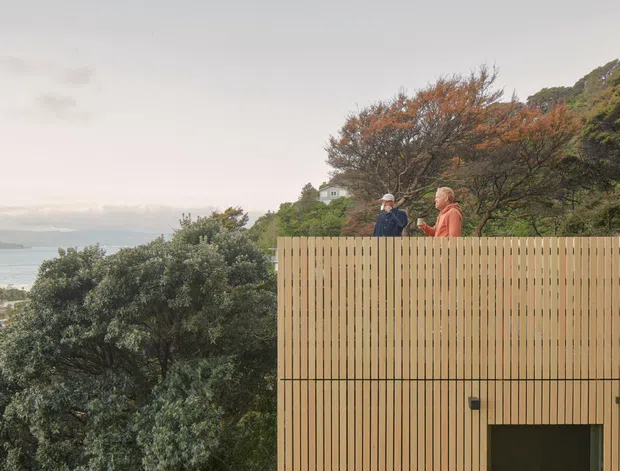 Exterior architectural view of a contemporary elevated building clad in vertical timber battens, with two people standing on a rooftop terrace or balcony. Dense coastal vegetation and a broad body of water are visible beyond the structure, with a small portion of a dark opening or doorway at the lower right.