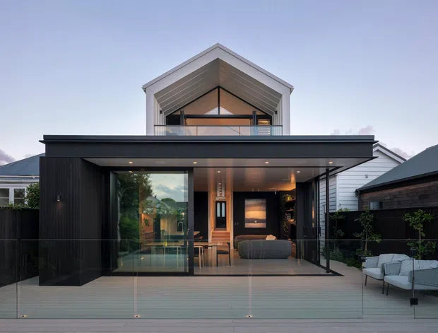 Contemporary two-storey residential extension viewed from the rear, featuring a dark flat-roofed lower level opening onto a broad timber deck and glazed balustrade. The upper level has a prominent gabled form with light cladding, exposed geometric roof lining and a balcony. Large sliding glazing reveals a warm-toned living area, while neighbouring residential buildings are visible on either side.