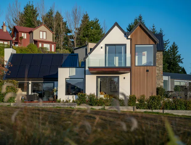 Exterior view of a contemporary multi-level house with steep gabled roof forms, white rendered walls, vertical timber cladding, large glazed openings, and a dark roof. Solar panels are mounted on a lower roof section, with landscaped planting around the building and neighbouring homes visible behind.