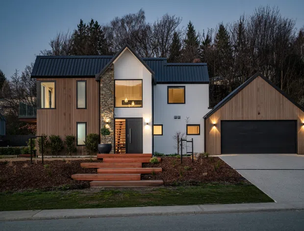 A contemporary two-storey residential home photographed at dusk, with a steep central gable, dark standing-seam metal roofs, mixed vertical timber and light rendered facades, and a prominent stone-clad entry feature. A separate matching gabled double garage sits to the right, connected by a concrete driveway. Warm interior and exterior lighting contrasts with the cool evening setting, while landscaped garden beds and timber entry steps frame the frontage.