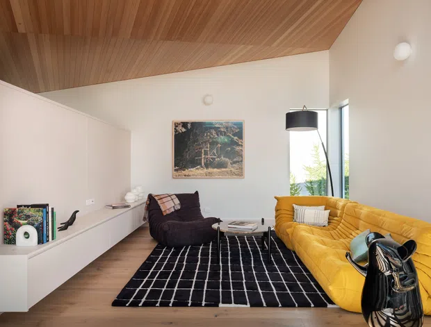 A stylish living room with a warm timber-lined ceiling, white walls, timber flooring, built-in white cabinetry, and large windows. The room features a mustard-yellow curved sofa, dark lounge chair, black-and-white grid rug, sculptural black side table or vase, pendant lighting, and framed artwork.