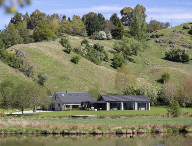 Wide exterior view of a low, elongated contemporary house set in a landscaped rural or semi-rural hillside site beside a calm pond or lake. The dwelling has dark grey stone walls, a dark pitched metal roof, extensive floor-to-ceiling glazing, and a connected low-profile wing. Rolling green terrain and mature trees form the backdrop.