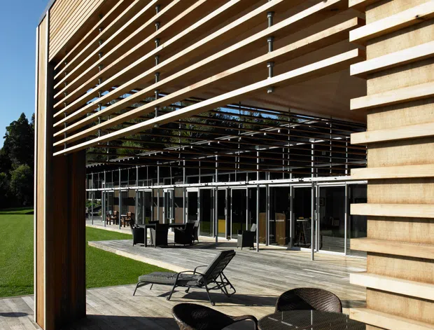 Exterior architectural view of a contemporary multi-level building with extensive timber horizontal slat screening and a deep covered outdoor terrace. Full-height glazing opens onto a timber deck furnished with dark woven outdoor chairs, loungers, and a round table. The open slatted facade creates strong linear shadows and passive shading, with landscaped lawn visible around the building.
