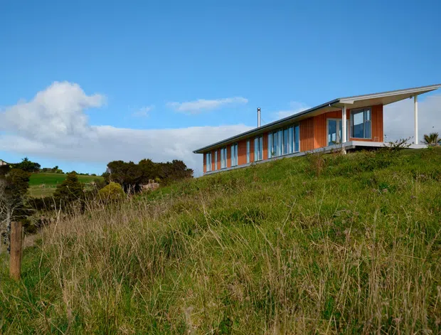 Exterior view of a long, low-slung contemporary house set on a grassy elevated or sloping site. The building features extensive vertical glazing, warm timber-coloured wall panels, a slim projecting roof with deep eaves, and a covered terrace or verandah at the right end. The surrounding rural or coastal-looking landscape is visible, with blue sky and grass dominating the foreground.