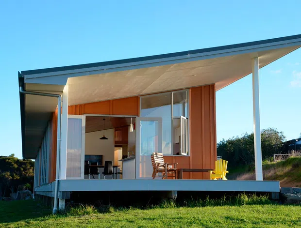 A compact modern single-storey pavilion-like house set in a grassy landscape. The building has warm vertical timber cladding, extensive sliding glass doors, a broad projecting roof with a slim dark edge, and a covered timber deck supported by slender posts. Interior furnishings are visible through the open glazing, including a dining area and chairs.