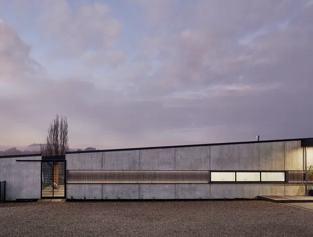A low, elongated contemporary building viewed externally at dusk, with a flat roof, dark grey concrete or masonry facade, long horizontal strip windows, and a recessed illuminated entry. A gravel forecourt and a waterfront or landscaped edge are visible in the foreground and distance.