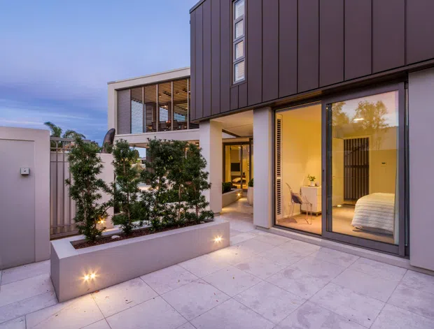 Contemporary multi-level residential facade viewed from a paved courtyard or terrace at dusk. The building combines dark vertical metal cladding with pale rendered walls, large floor-to-ceiling glazed sliding doors, narrow vertical windows, and a recessed illuminated entry. A bedroom is clearly visible through the glazing, while a raised planter with small trees and integrated outdoor lights defines the courtyard edge.