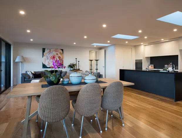 Open-plan contemporary kitchen, dining and living area with warm timber flooring, a large dining table, upholstered dining chairs, dark kitchen island cabinetry, white walls and ceiling, recessed lighting, and multiple skylights. Floor-to-ceiling glazing lines the left side, while a lounge area and decorative built-in shelving are visible beyond the dining zone.