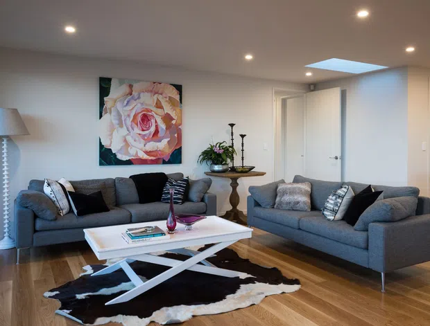 A bright, contemporary living room with two charcoal-grey sofas arranged around a white rectangular coffee table. The room features warm timber flooring, white walls and ceiling with recessed downlights, a large floral artwork, a sculptural floor lamp, decorative console table, patterned cushions, and a black-and-white cowhide rug. A roof skylight adds natural light, and doorways are visible at the rear.
